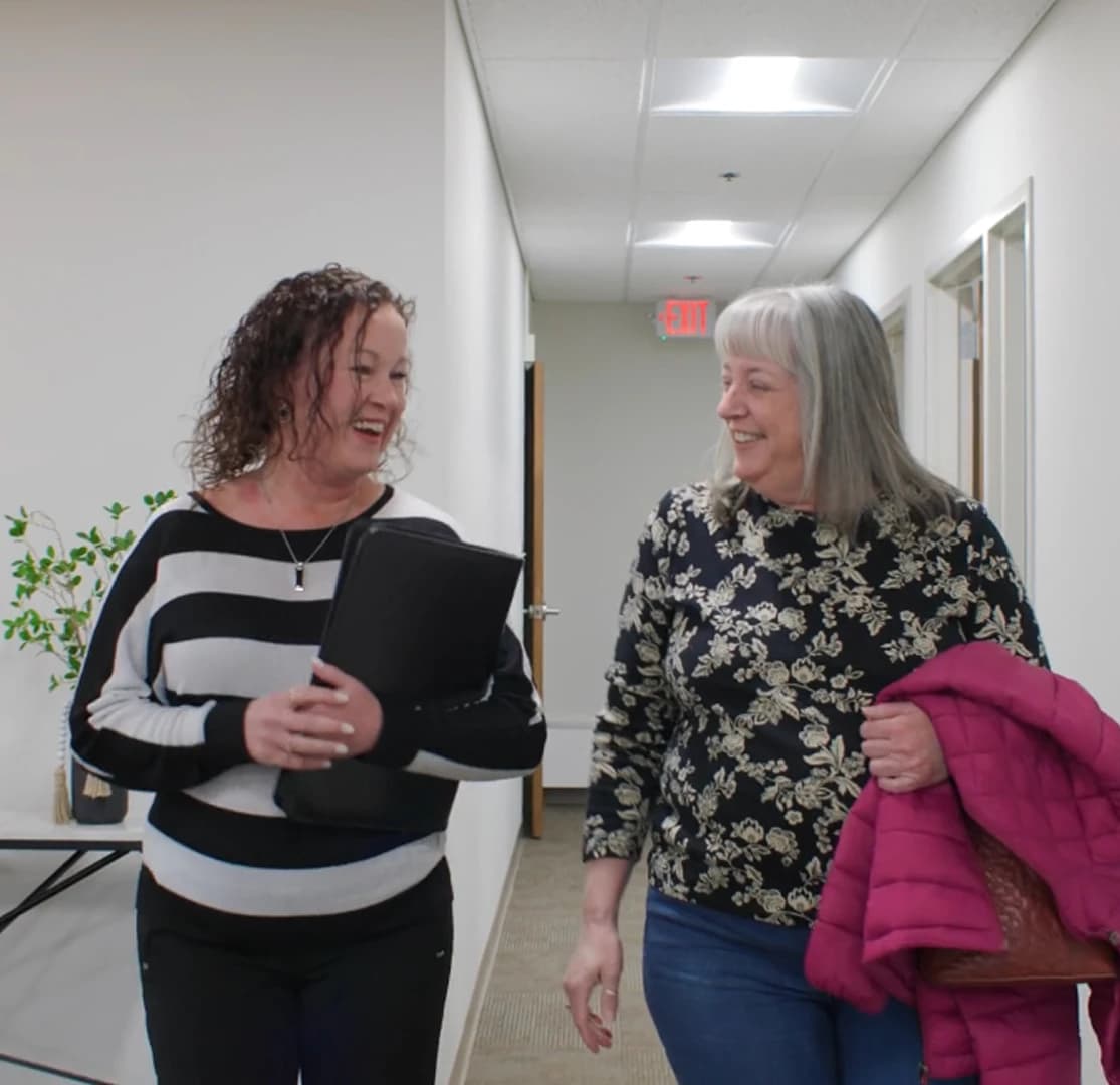 Two smiling women walking together through a bank office hallway